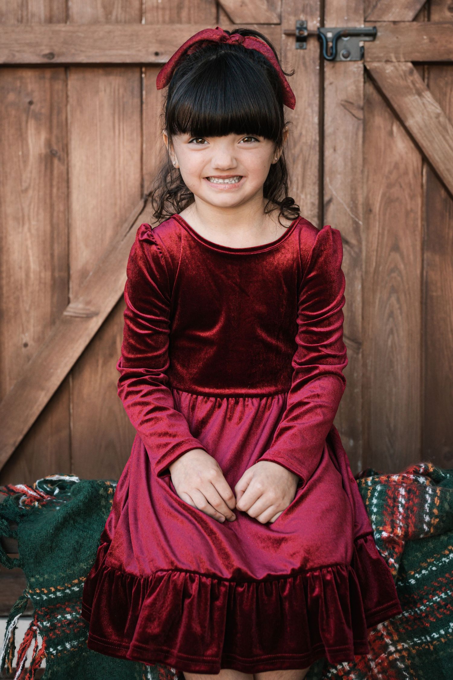 Young girl in burgundy velvet long-sleeve dress with ruffle hem and red bow headband posing before wooden barn doors.