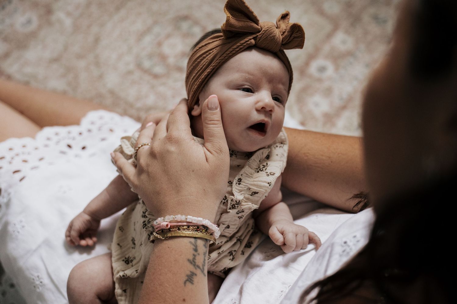 Mother with gold bracelet and tattoo gently holds newborn baby wearing brown bow headband and floral outfit.