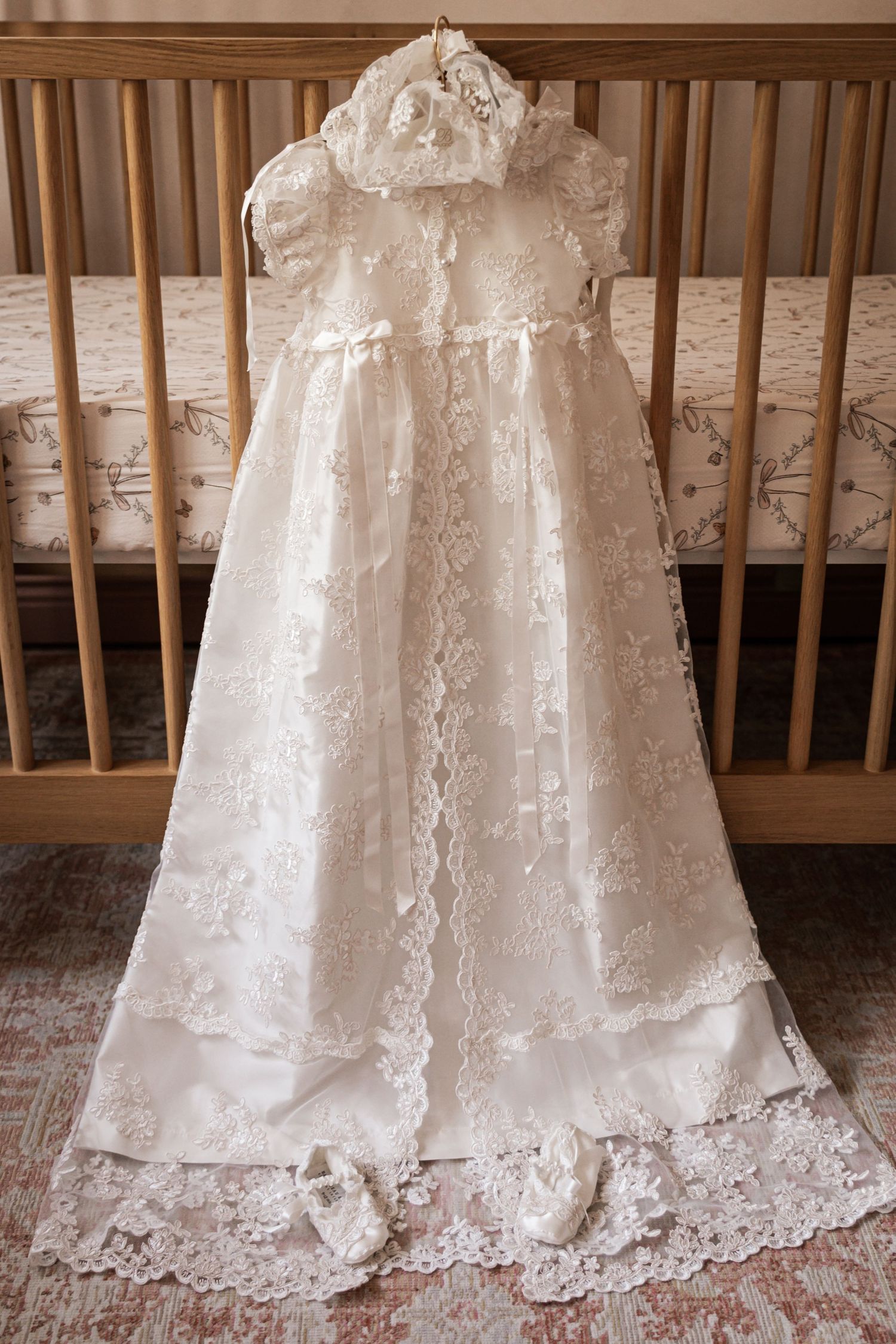 A beautiful white lace christening gown with matching bonnet and shoes displayed on a wooden baby crib.