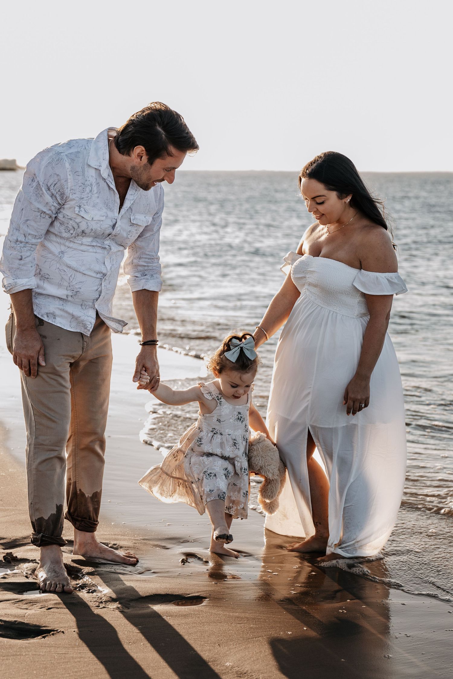Happy family with pregnant mother and toddler daughter playing at beach shoreline.