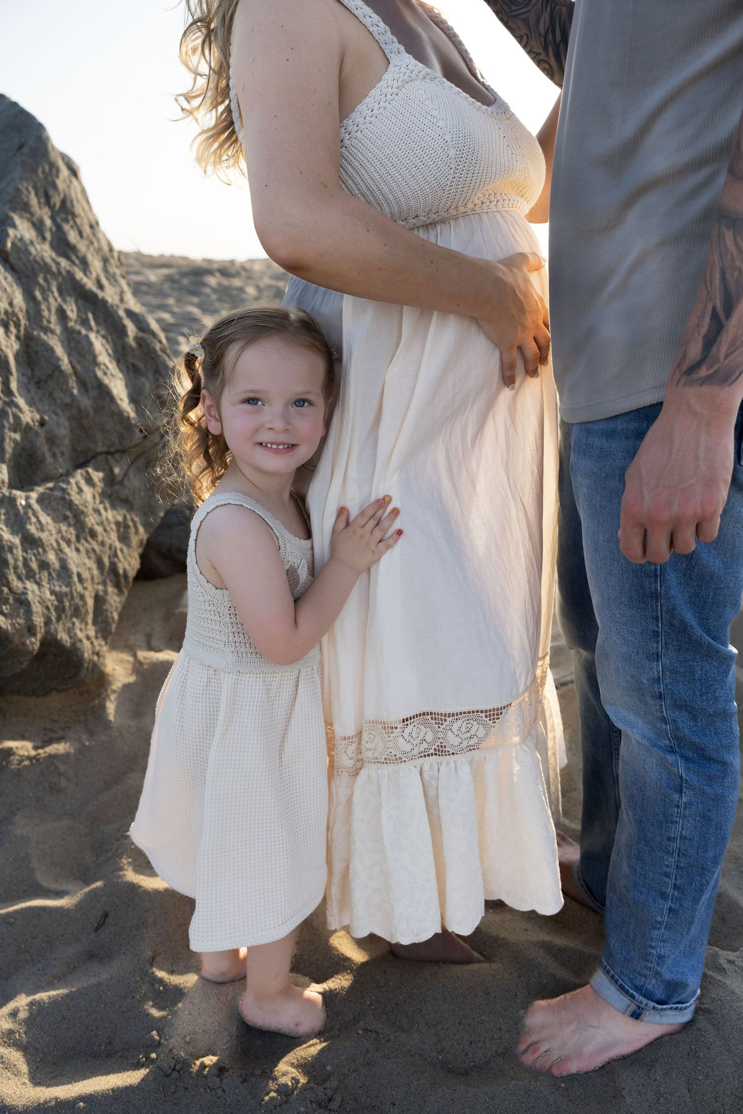 Smiling toddler girl in white dress hugging pregnant mothers skirt at beach.