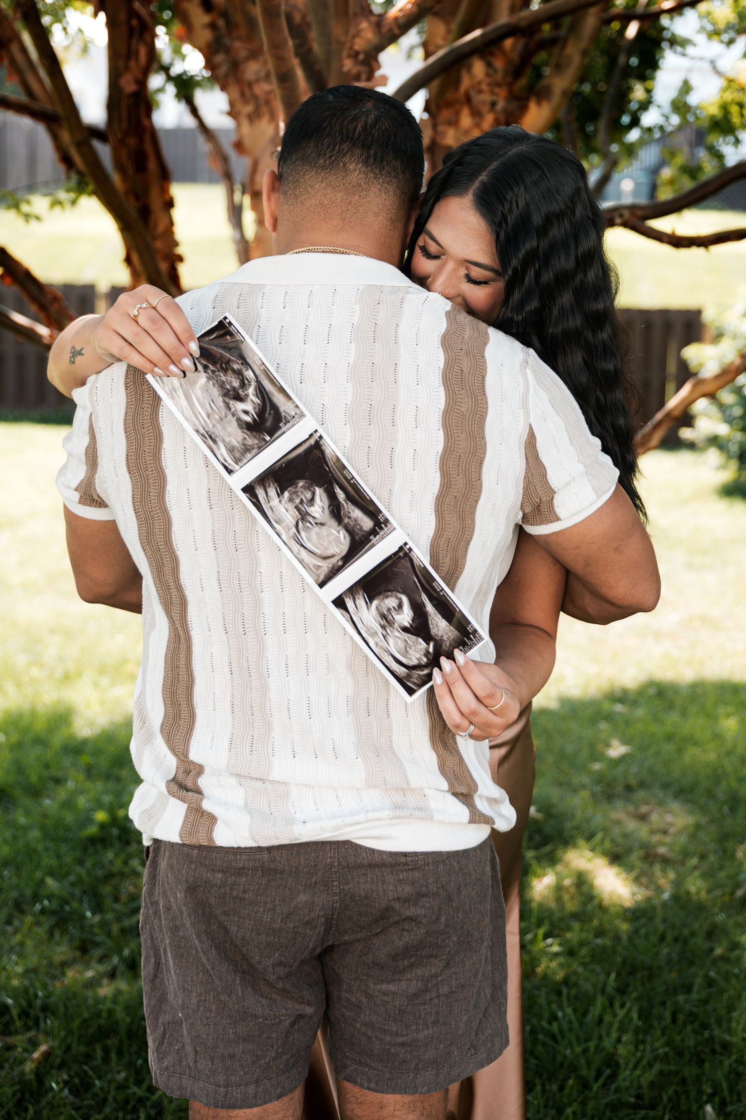 Couple embracing outdoors holding ultrasound photos, pregnancy announcement.