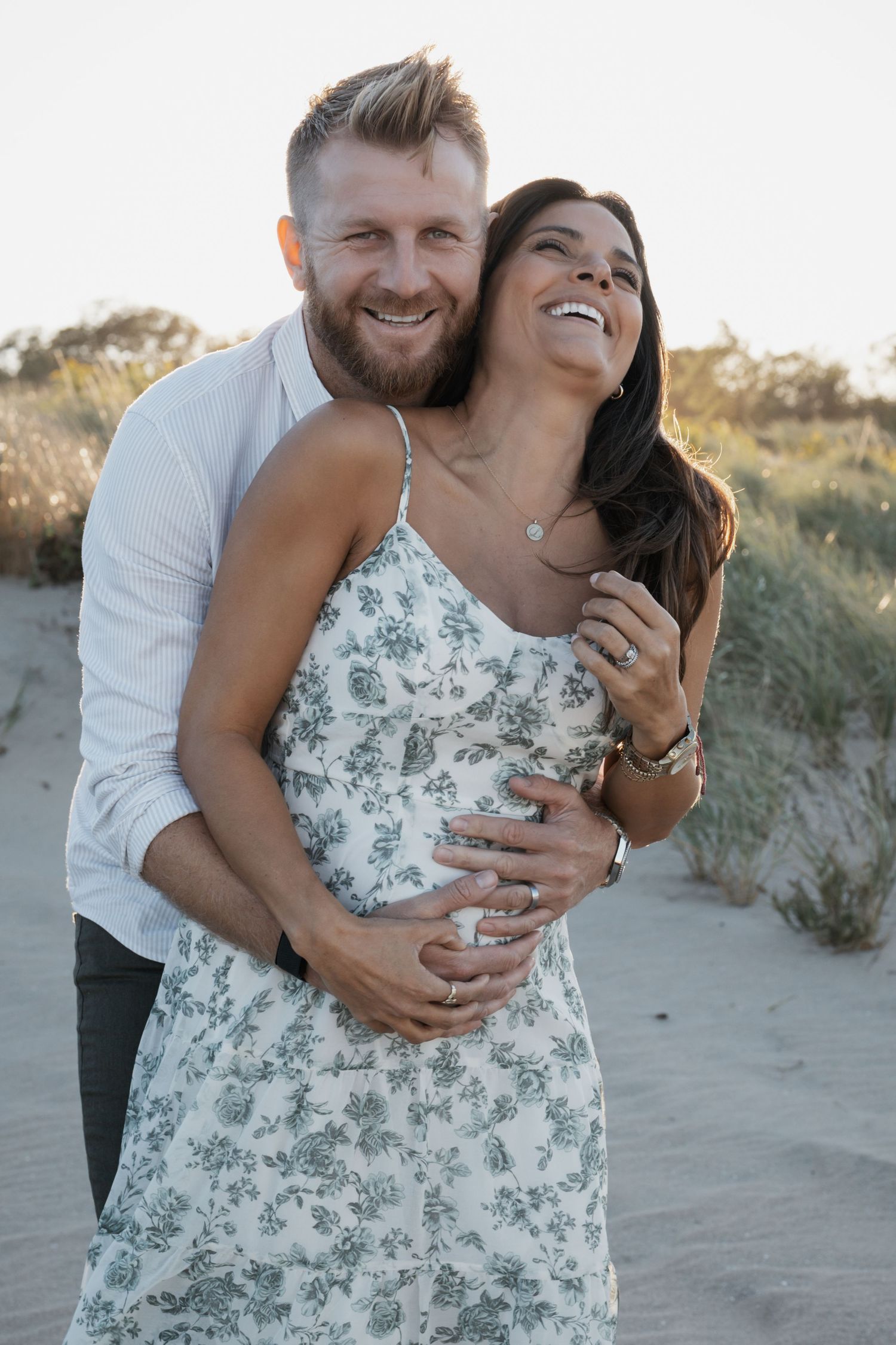 Bearded man embracing laughing pregnant woman in floral dress on sandy beach at sunset.