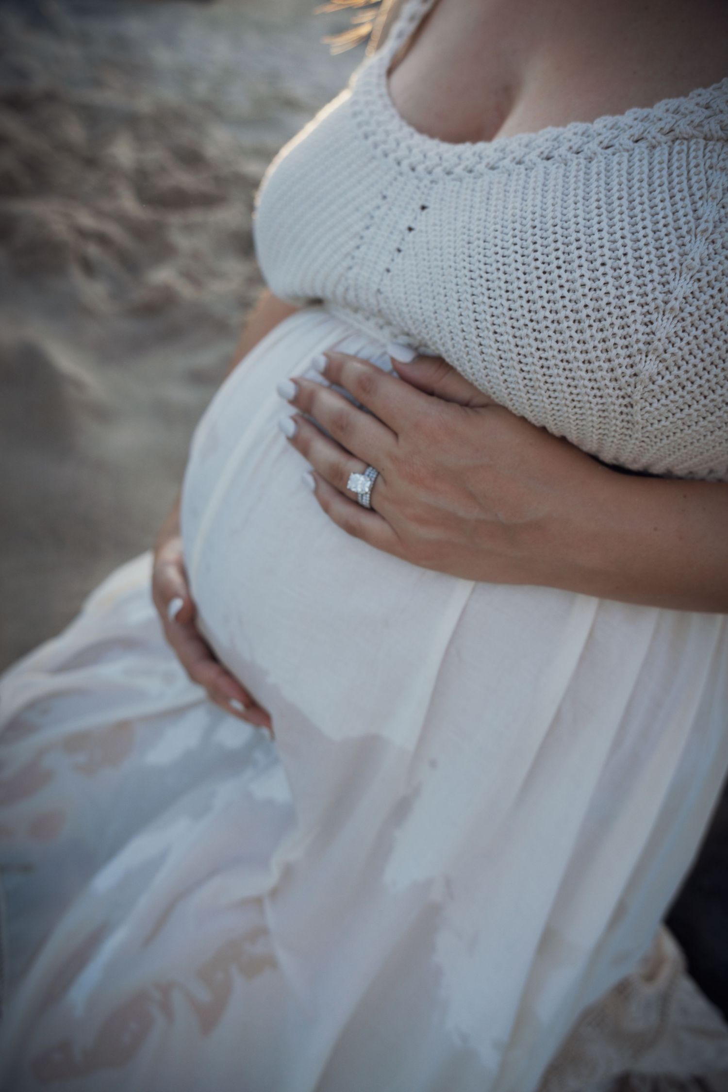 Close-up of pregnant belly with diamond ring, white maternity dress on beach.