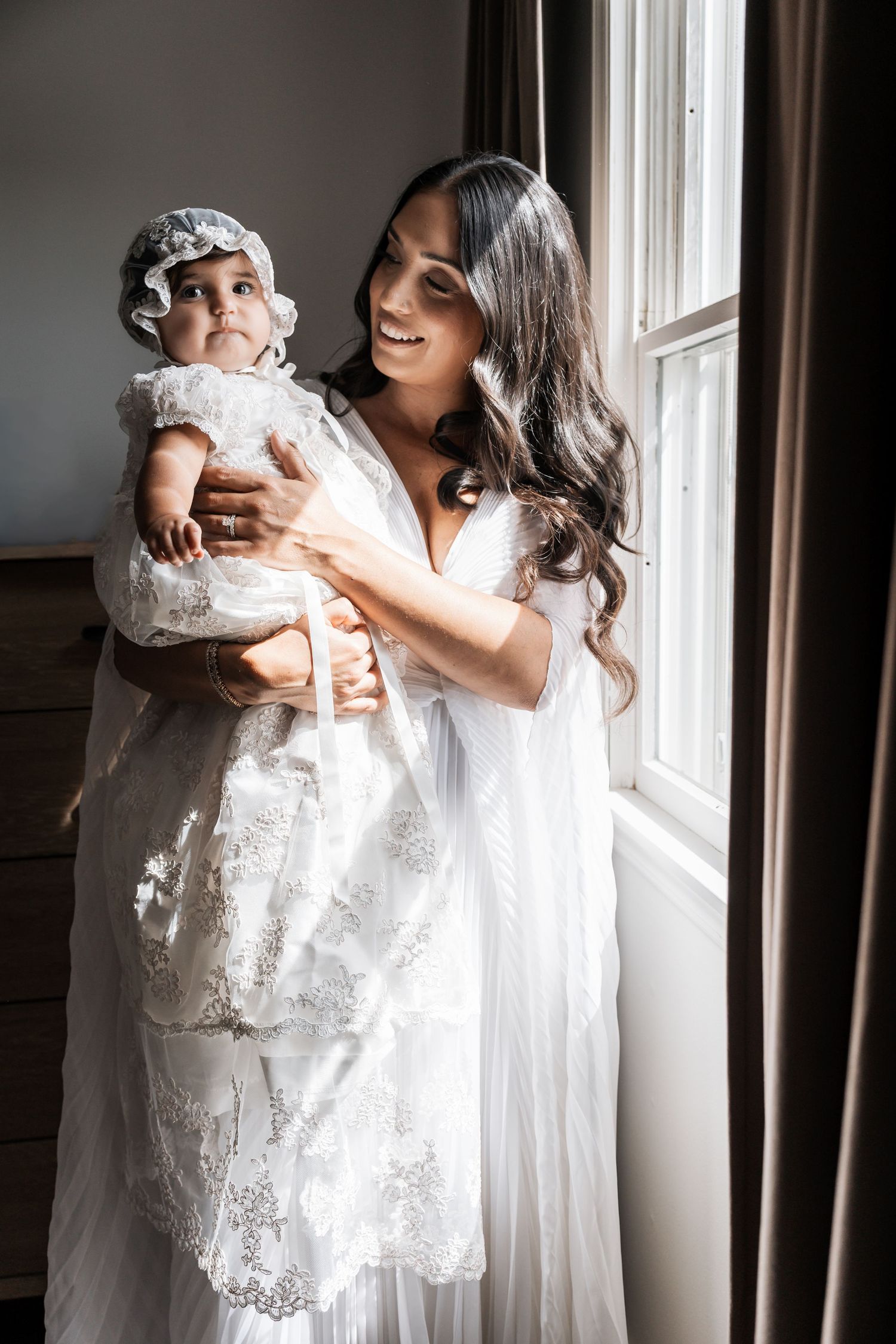 A smiling dark-haired mother in white dress holds her baby in a lace christening gown by a sunlit window.