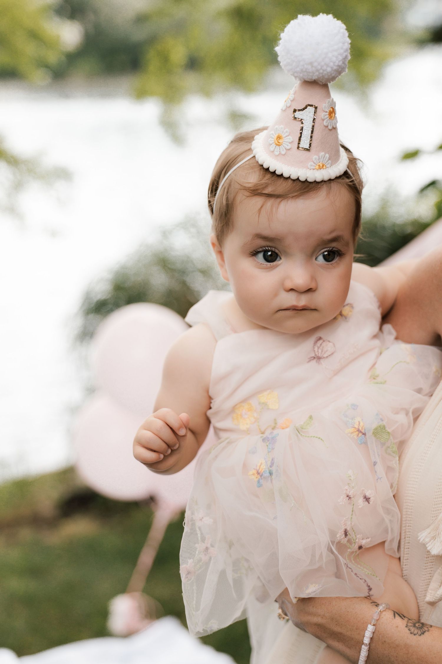 Baby girl wearing number one birthday hat and pink floral dress near balloons.