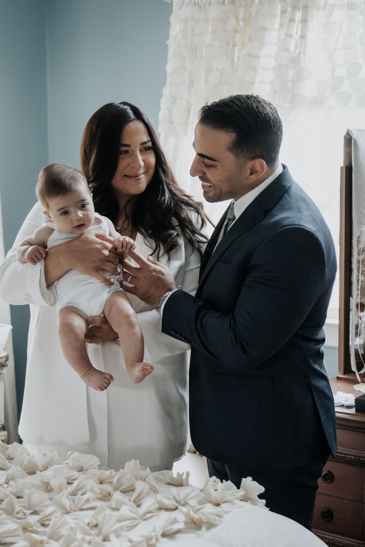 Elegant couple in formal wear holding baby in white christening gown beside decorated bed.