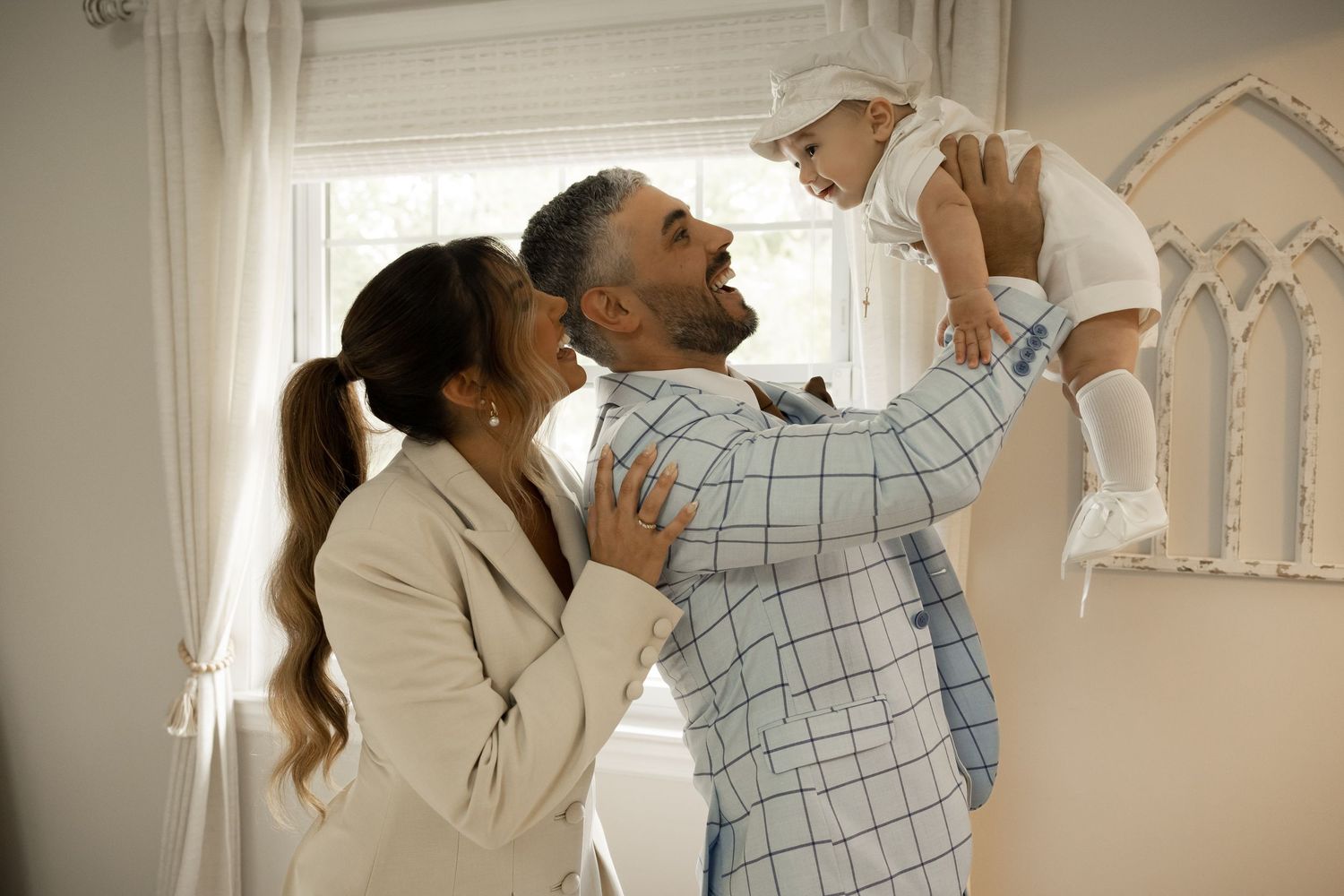 Happy parents in white outfits lifting baby in white christening clothes in bright nursery.