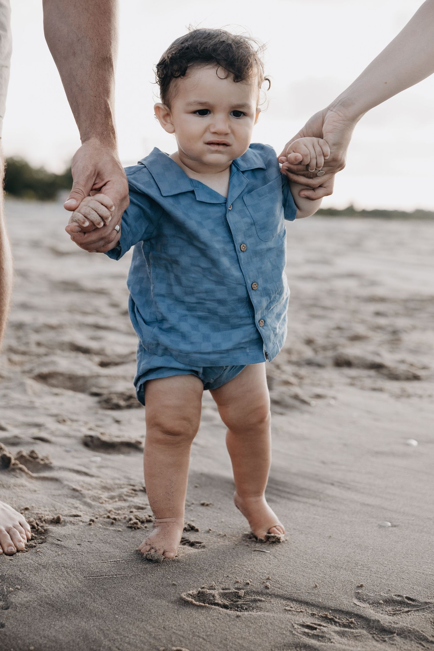 Toddler in blue outfit taking first steps on sandy beach with parents.
