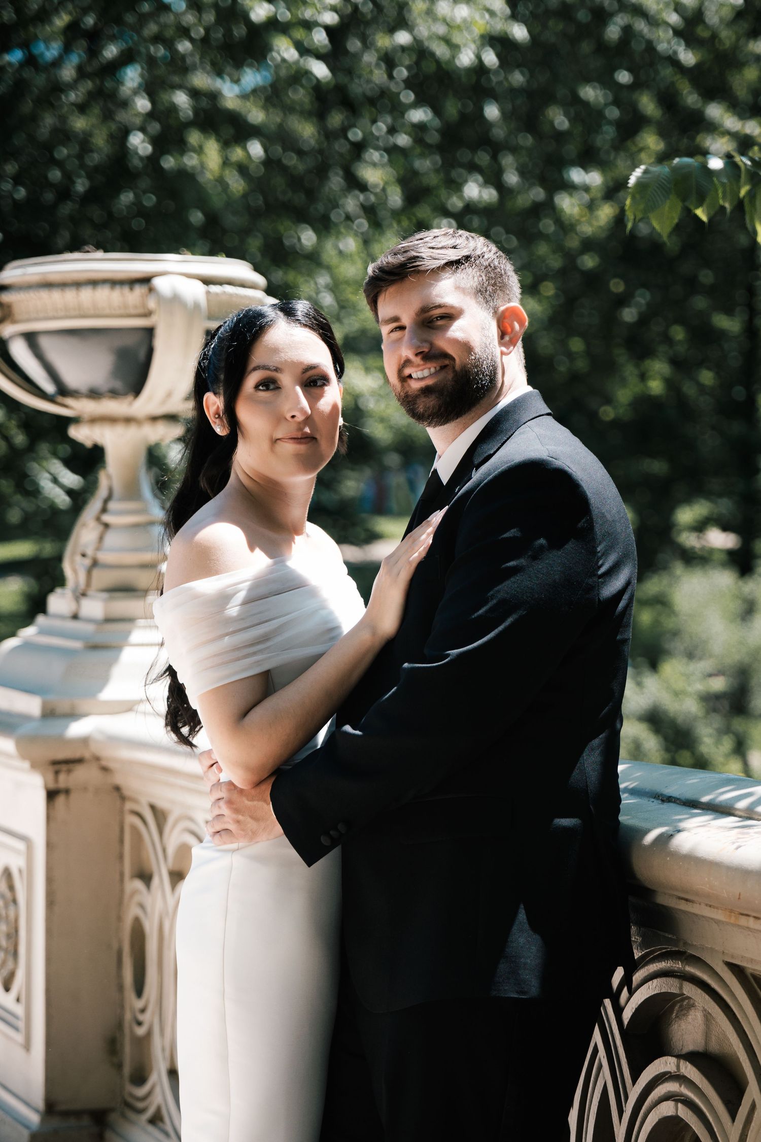 Elegant bride in off-shoulder white gown and bearded groom in black tuxedo pose at stone balustrade in New Jersey.