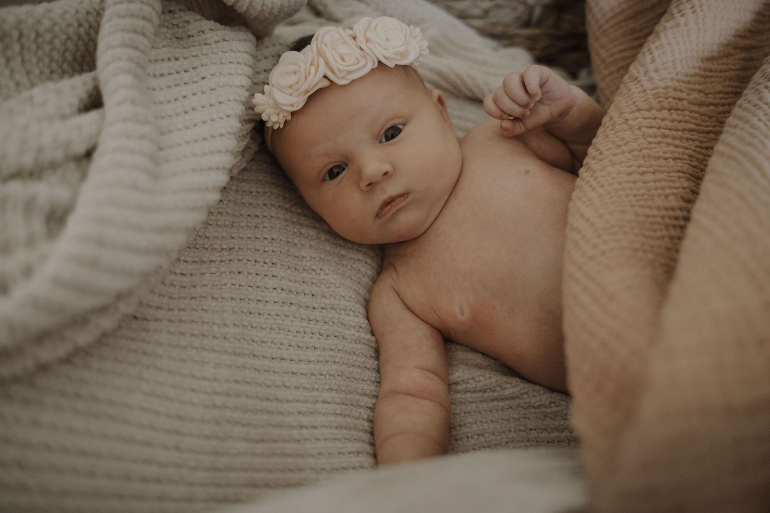 Newborn baby girl with floral headband lying on a soft beige knit blanket.