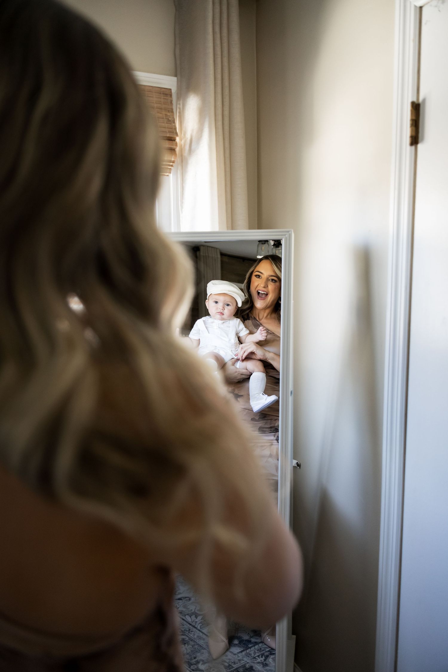 A smiling mother holds her baby in white baptism outfit, reflected in a full-length mirror.