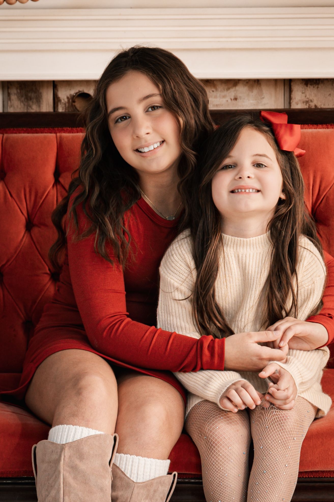 Two sisters in red and cream dresses sit closely together smiling on a red velvet sofa.