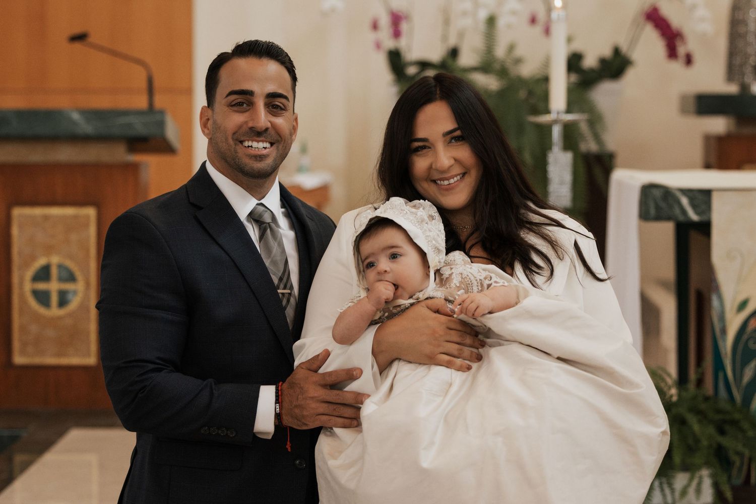Smiling couple holding baby in white christening gown and bonnet inside a church.
