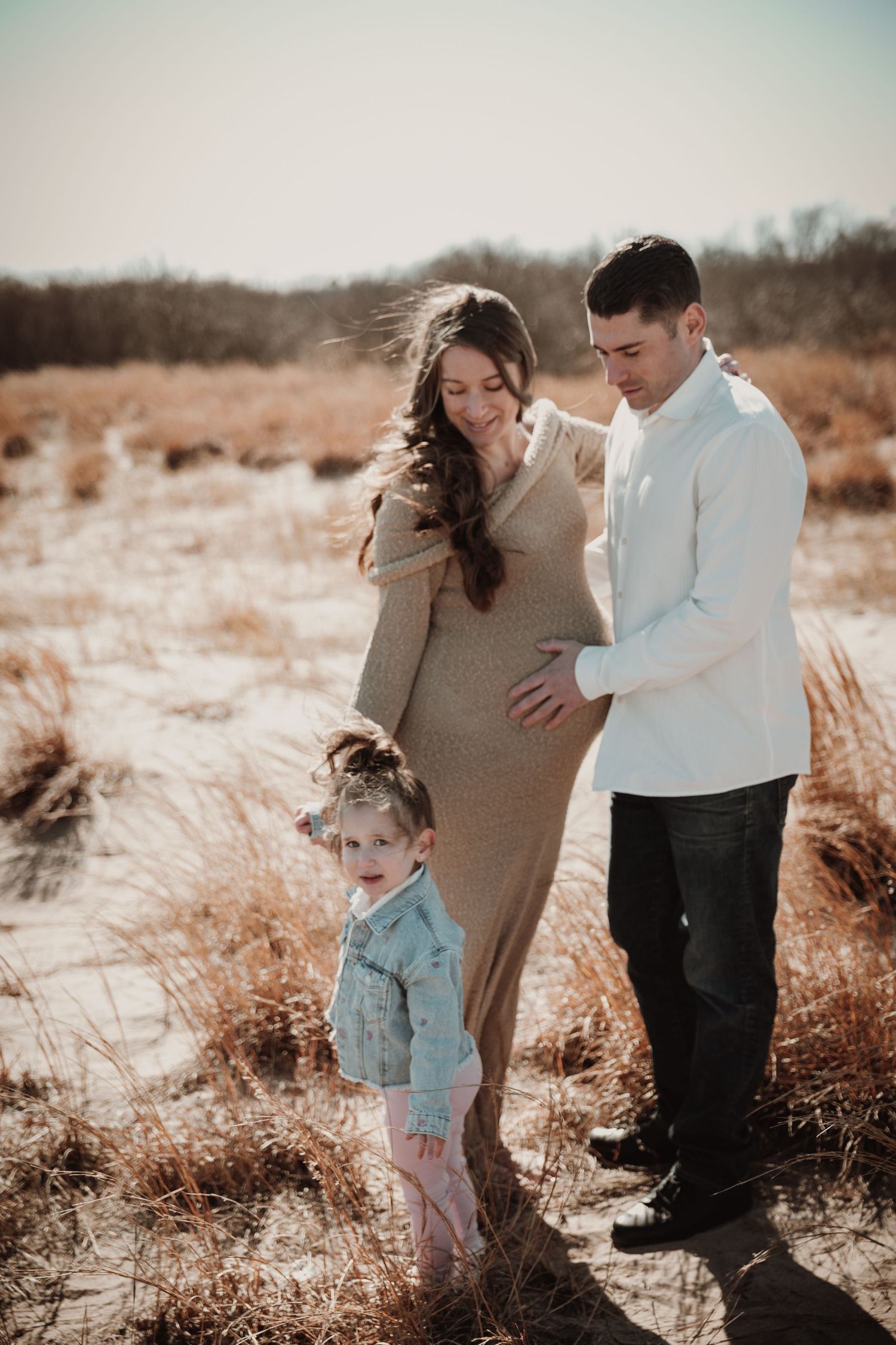 Expecting couple with young daughter in golden field during outdoor maternity photography session.