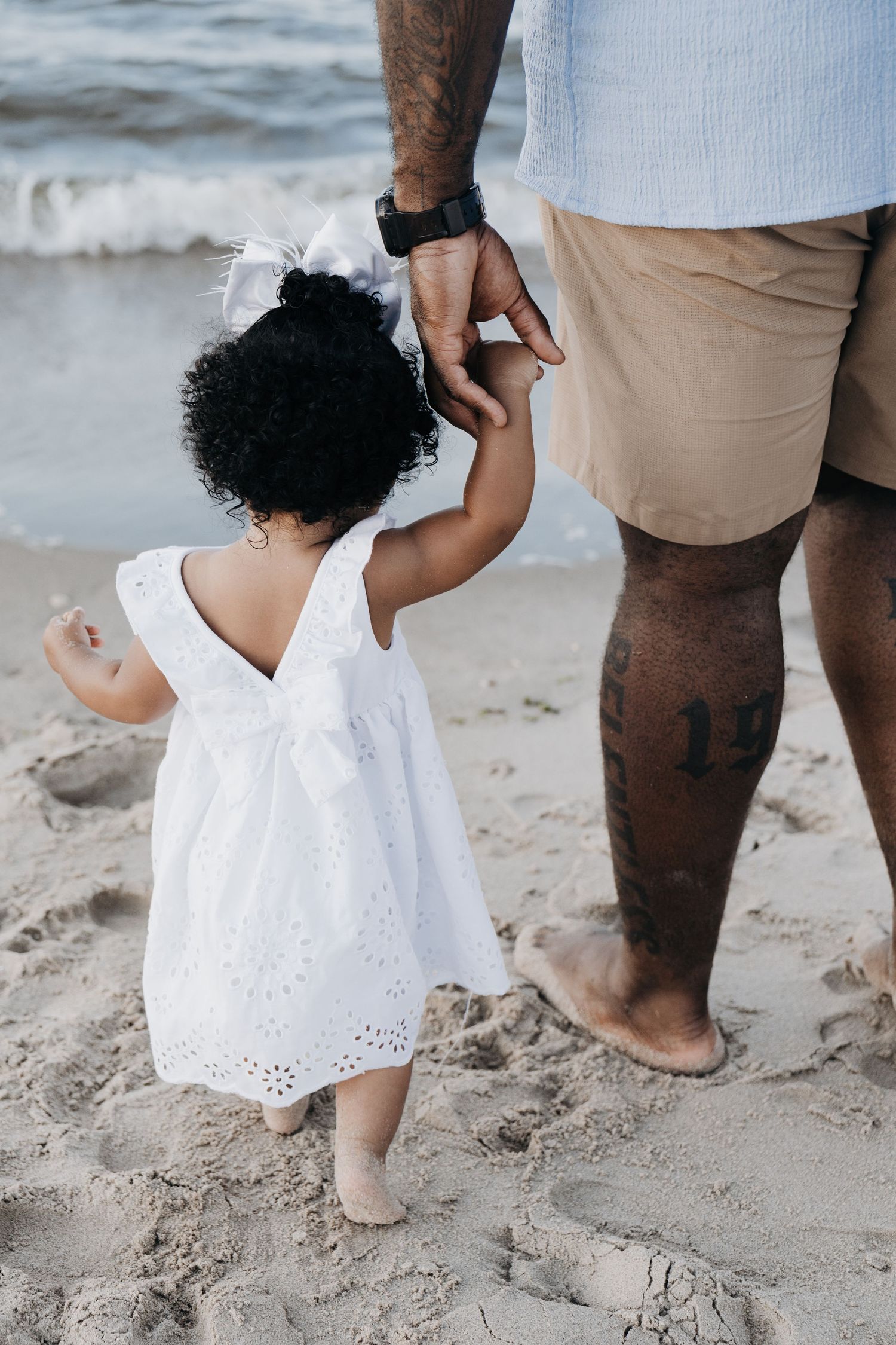 A toddler girl in a white dress holds her father's hand on a sandy beach near the ocean waves.