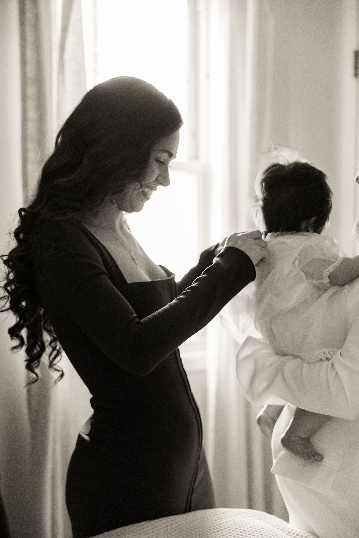 Black and white photo of mother smiling at baby in white christening gown by bright window.
