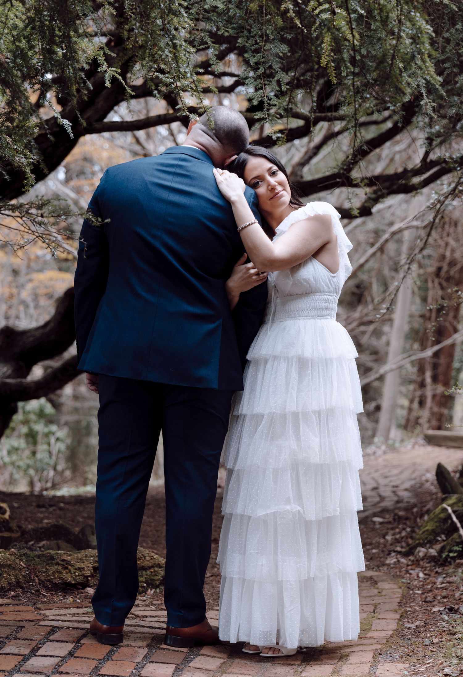 Bride in tiered white dress embracing groom in navy suit under scenic garden tree in New Jersey.