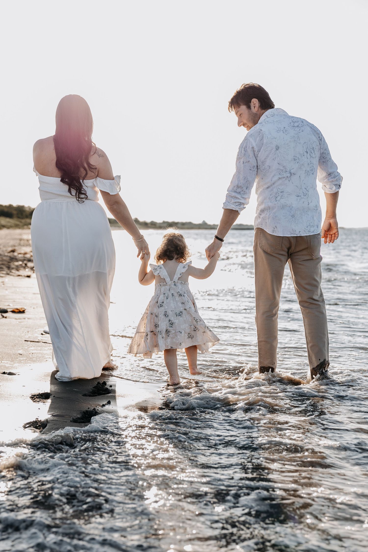 Family walking in ocean waves at sunset, parents holding toddler's hands.