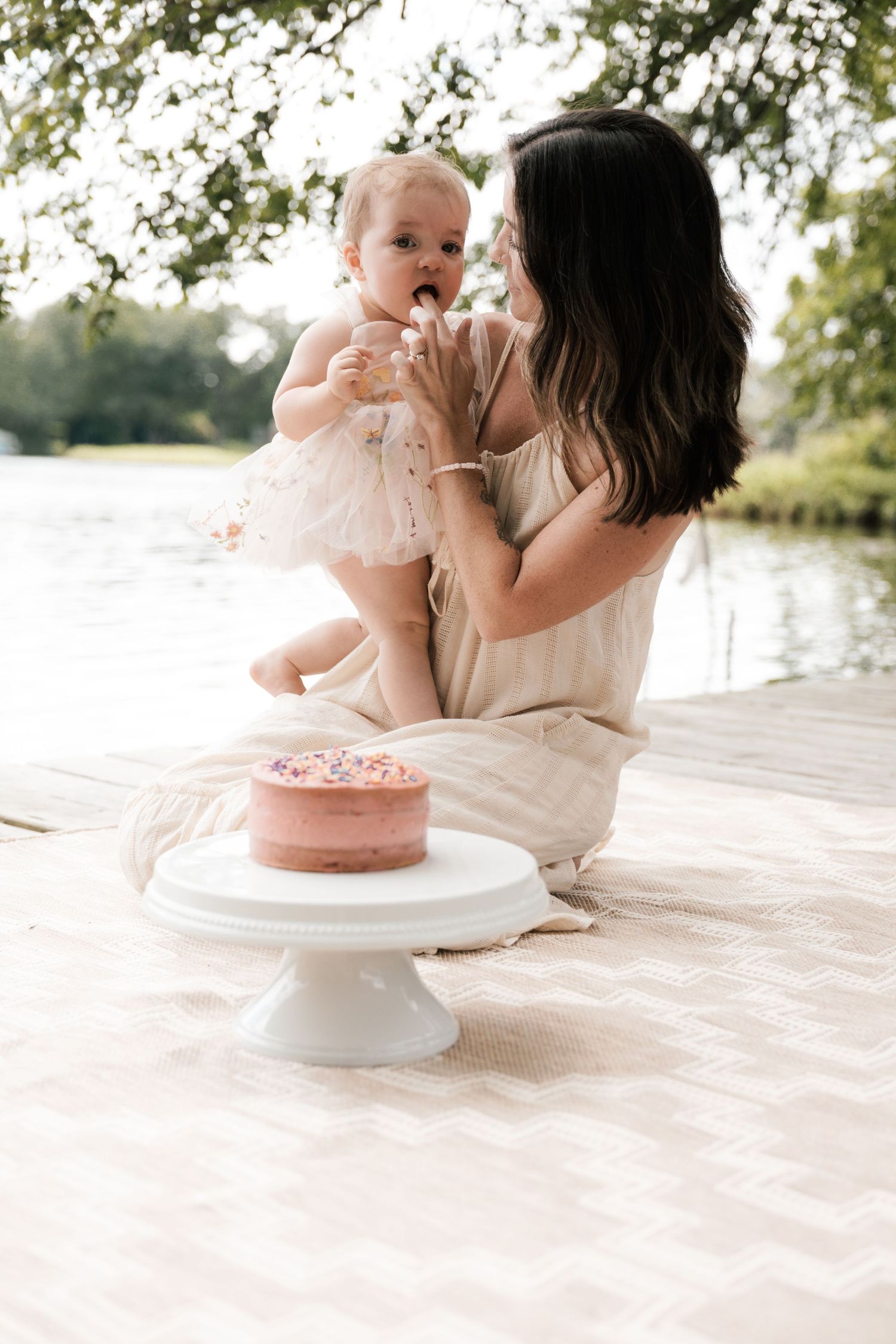 Mother holds baby girl near pink birthday cake on white cake stand by lake.