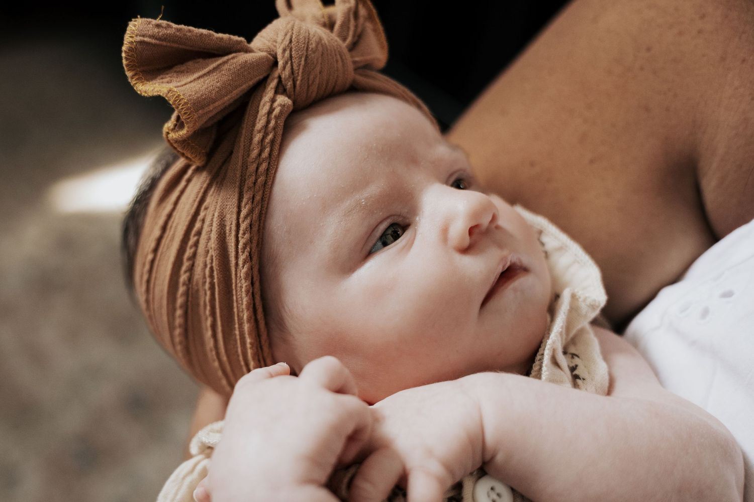 Peaceful newborn baby with brown knit bow headband resting in parents arms with hands folded.