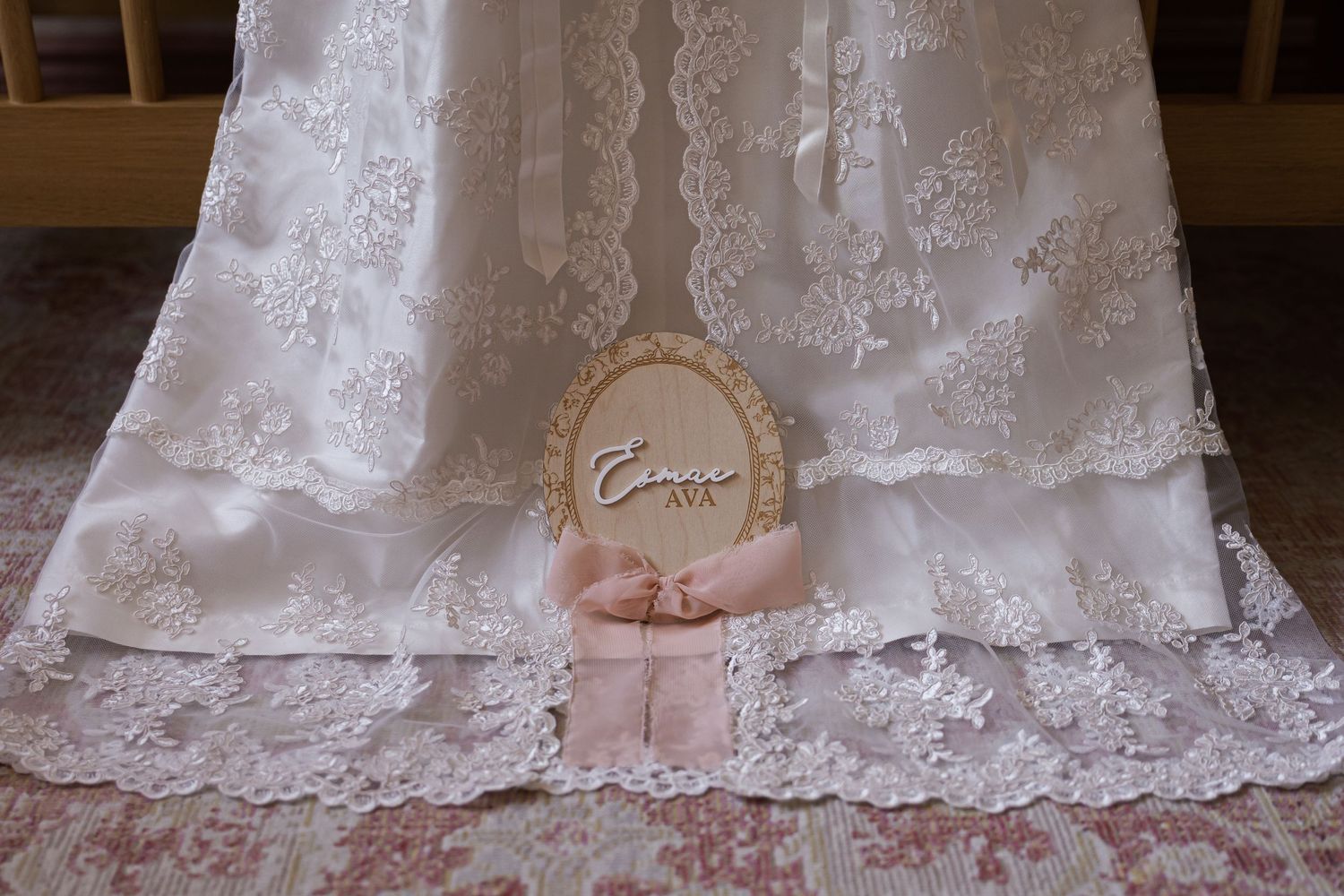 Close-up of an elegant white lace christening gown with a personalized wooden name tag and pink ribbon.