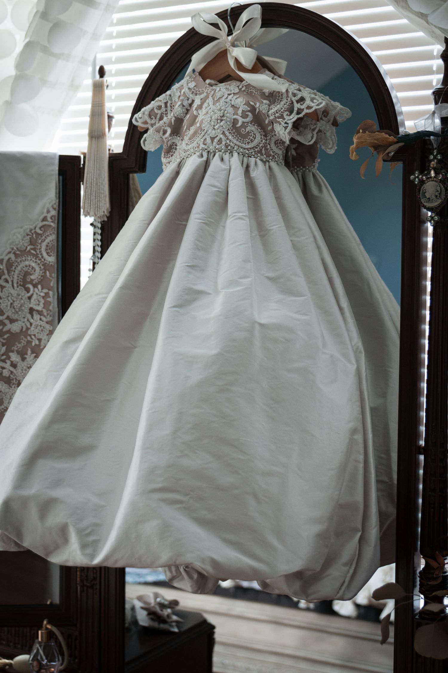 Delicate white baby christening gown with lace bodice hanging in a nursery room.