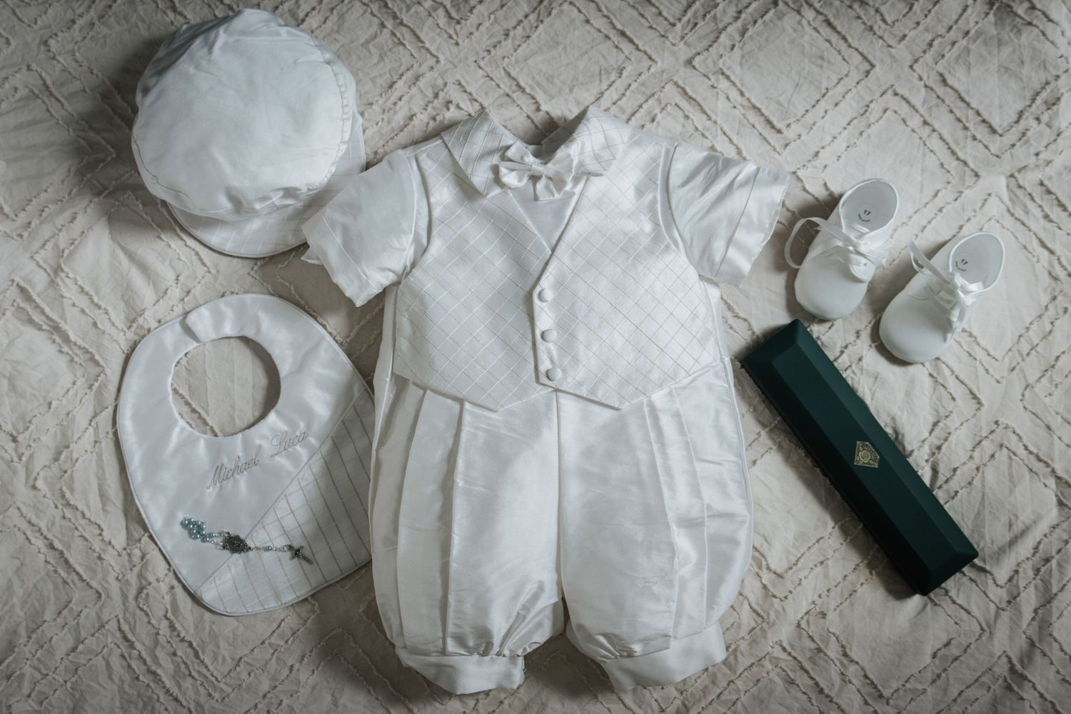 Flat lay of white baby christening outfit with hat, bib, shoes, and green book.