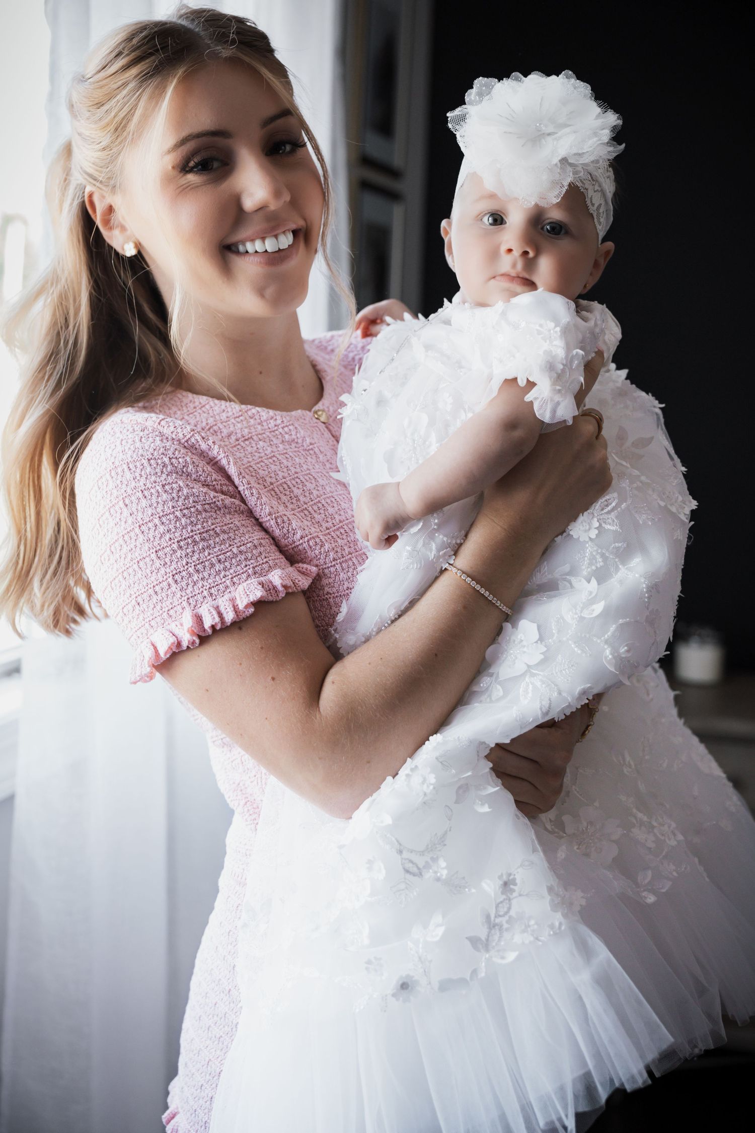 Cheerful blonde woman in pink tweed top holding baby in white lace christening gown with floral headband.