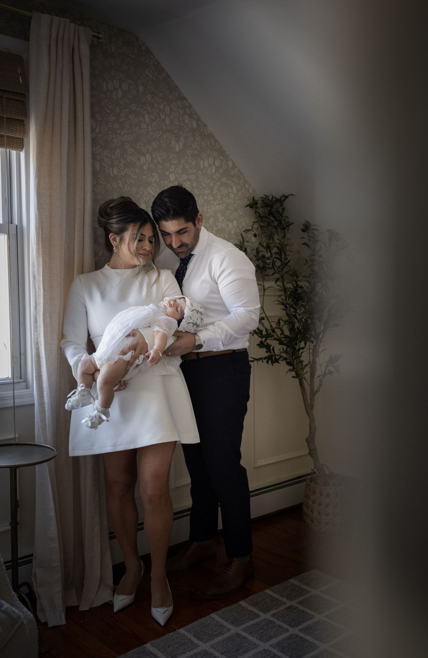 Elegant couple in white and black attire tenderly holds their newborn in a christening gown.