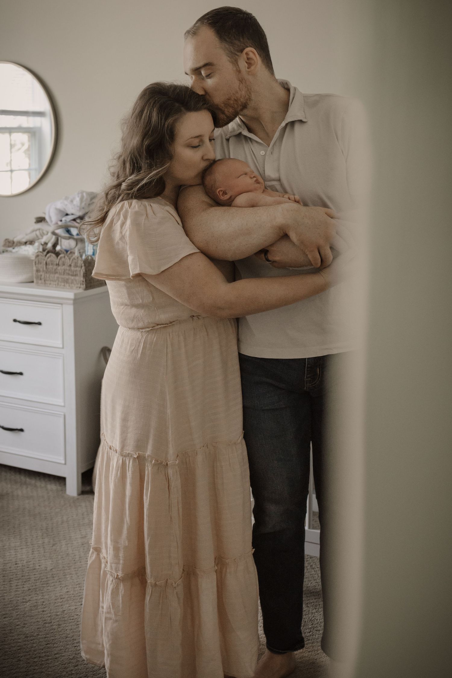 New parents embrace their sleeping newborn baby together in a softly lit nursery.