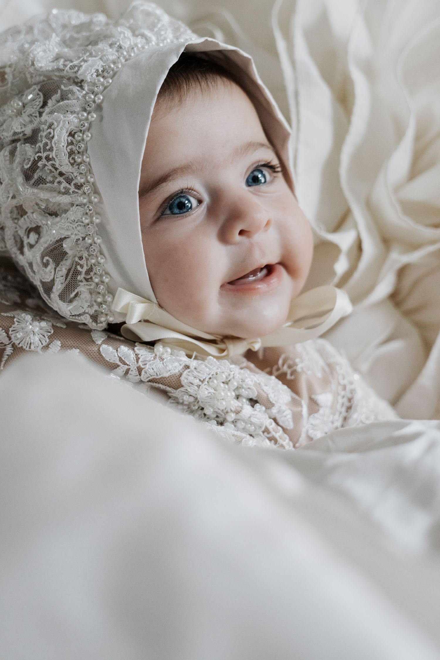 Close-up of blue-eyed baby smiling in detailed lace christening bonnet and white gown.