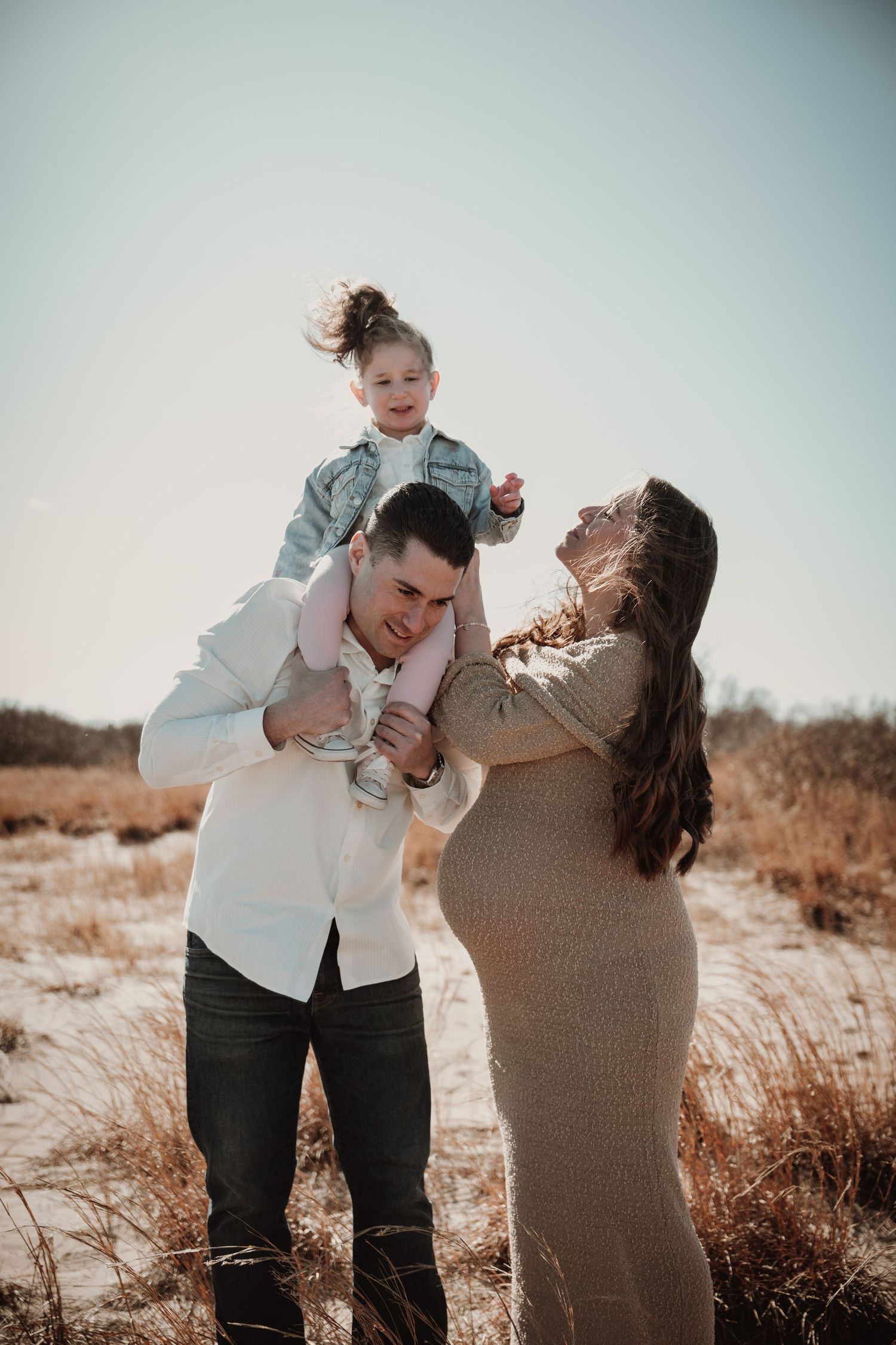 Happy family maternity photo with dad holding toddler on shoulders while pregnant mom laughs outdoors.