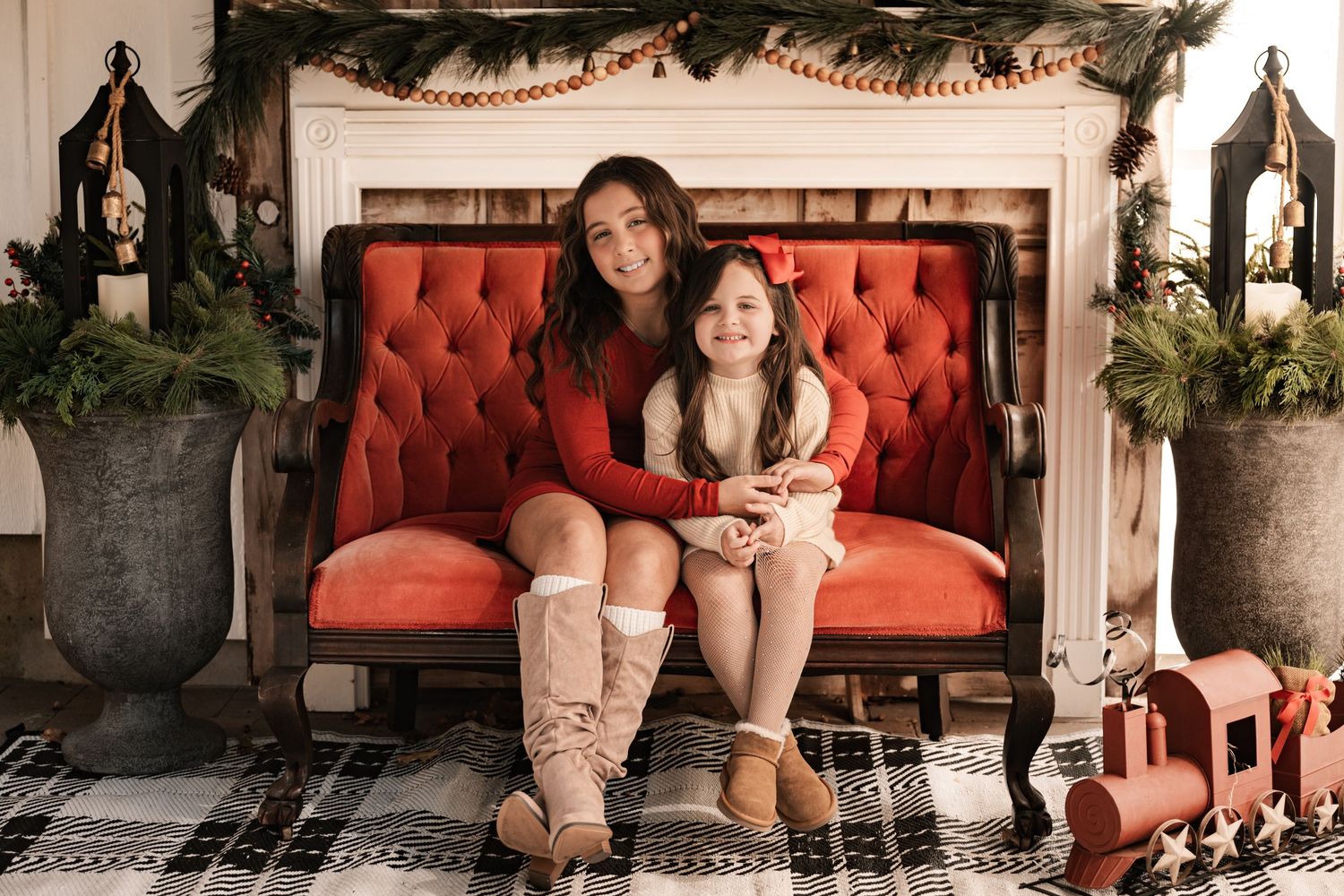 Two sisters smile together on a red velvet sofa in front of a Christmas fireplace decorated with greenery.