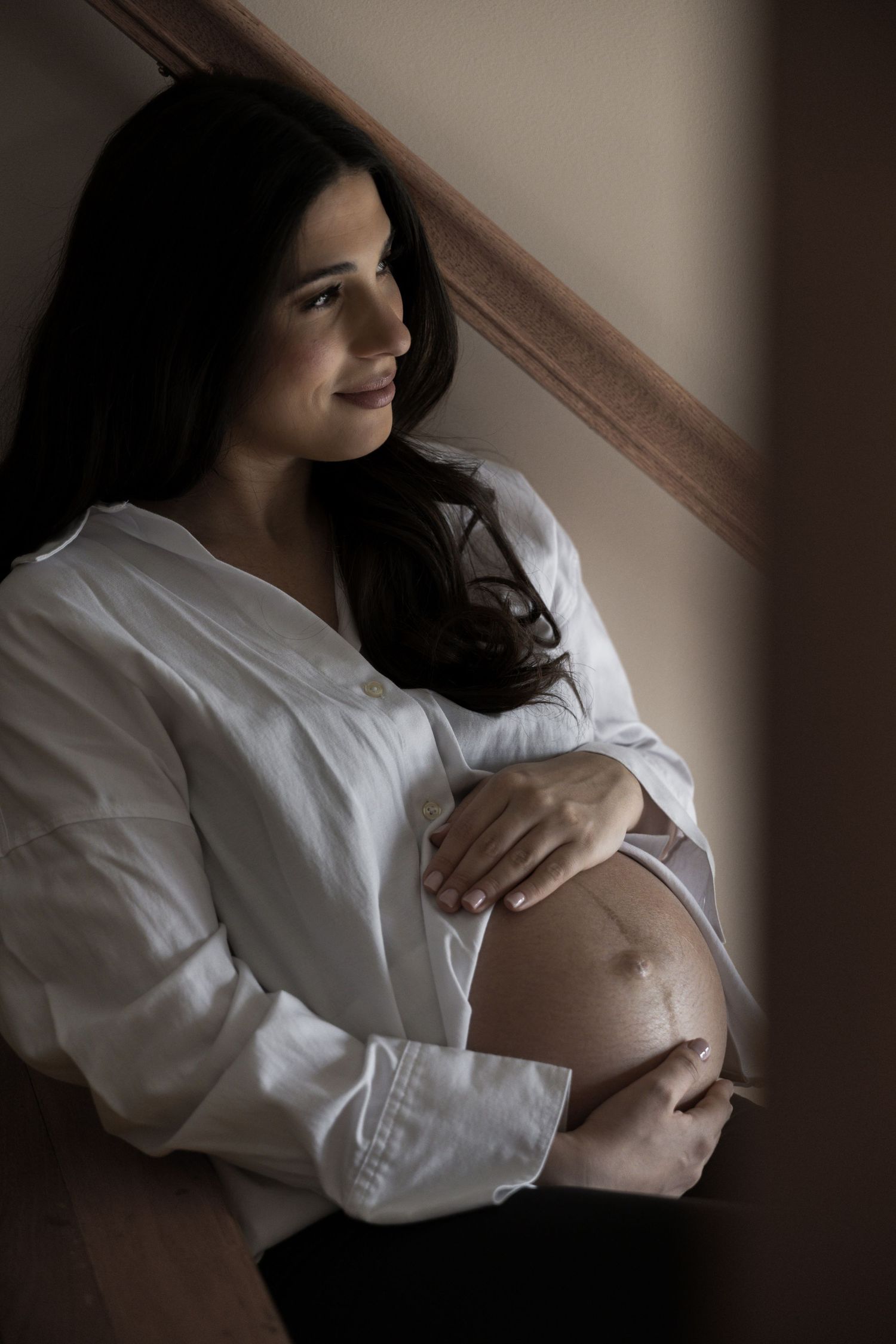 Pregnant woman in white shirt cradling bare baby bump while leaning against staircase indoors.