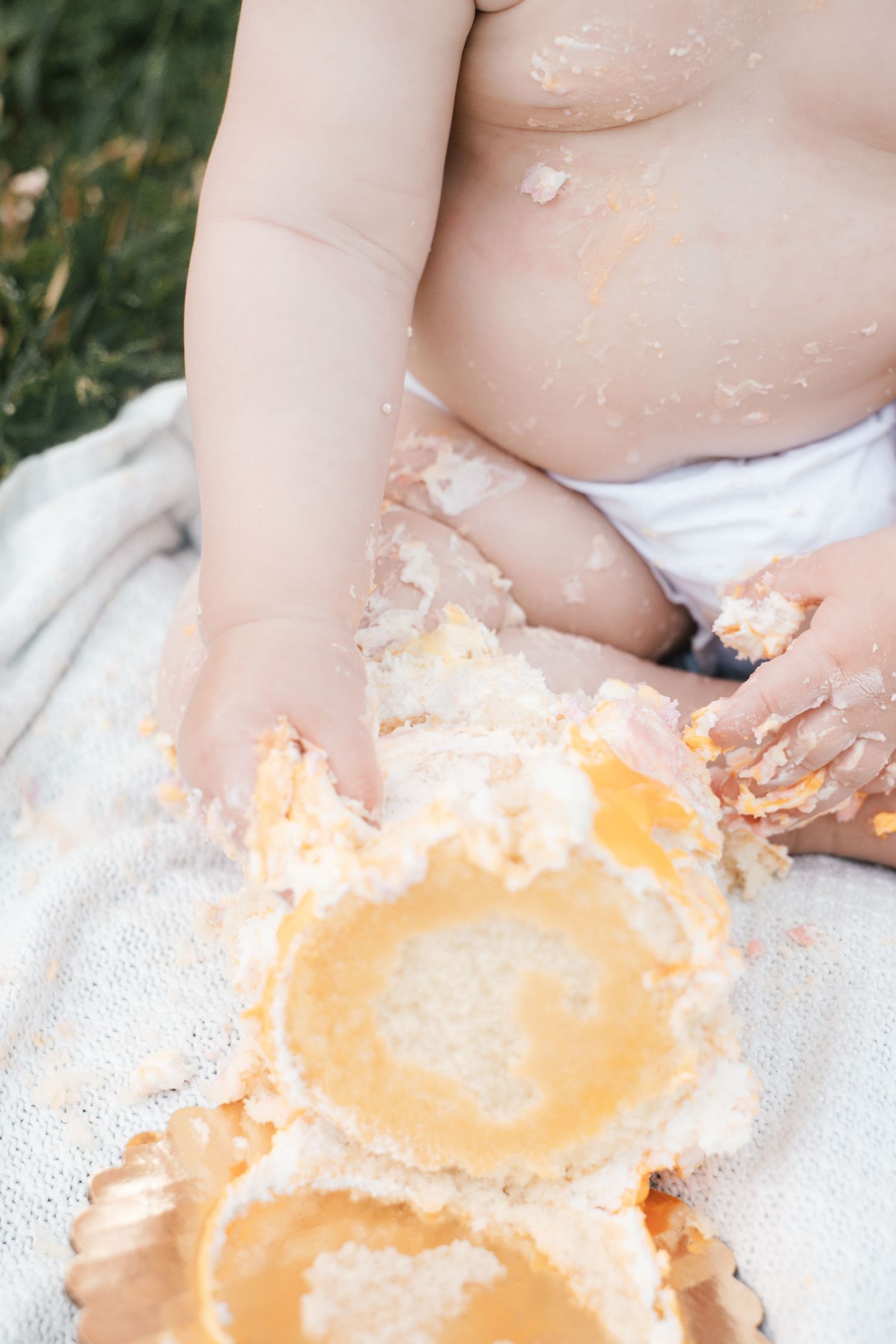 Baby enjoying first birthday cake smash outdoors, covered in white and yellow cake frosting.