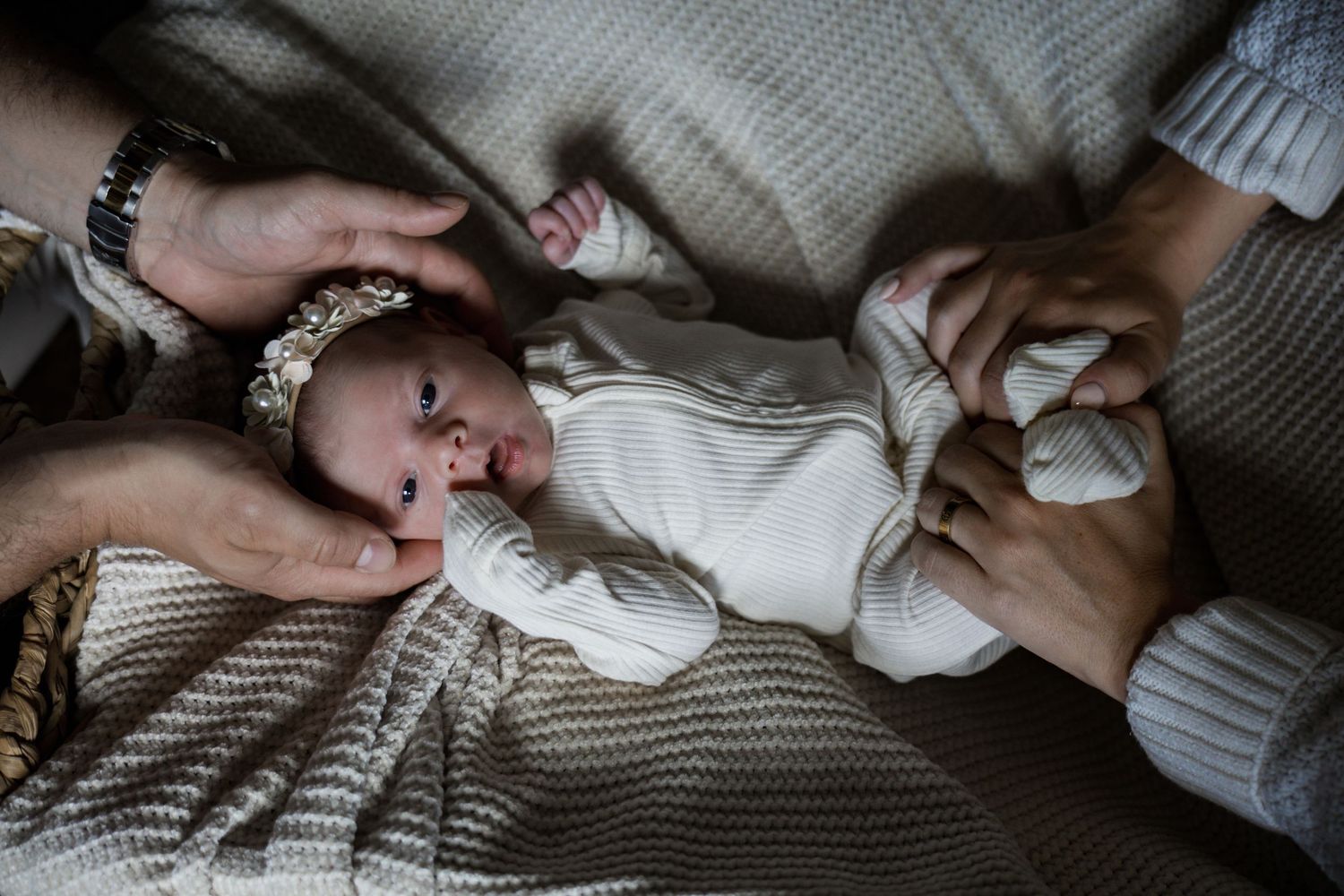 Parents hands tenderly cradle newborn baby in white onesie on a knit blanket.