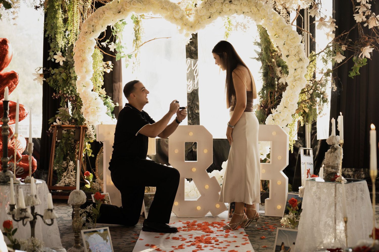 Man proposing to woman under a floral heart arch surrounded by romantic rose petal decorations.
