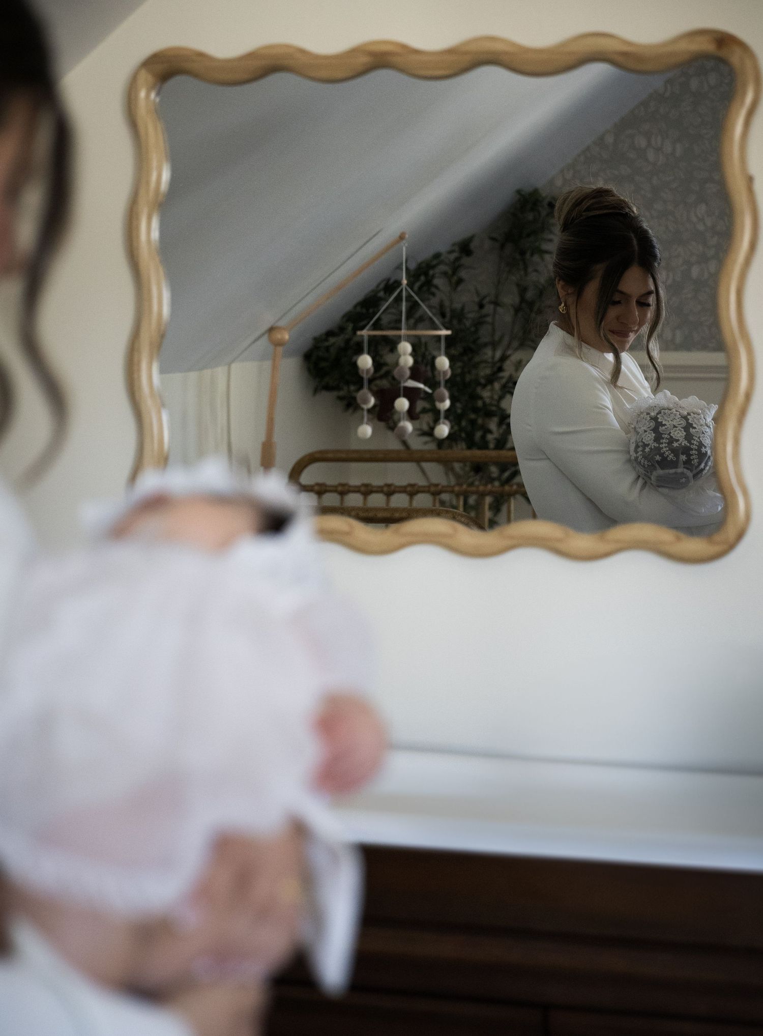 Mother holding baby in white christening gown reflected in ornate gold-framed mirror in nursery room.