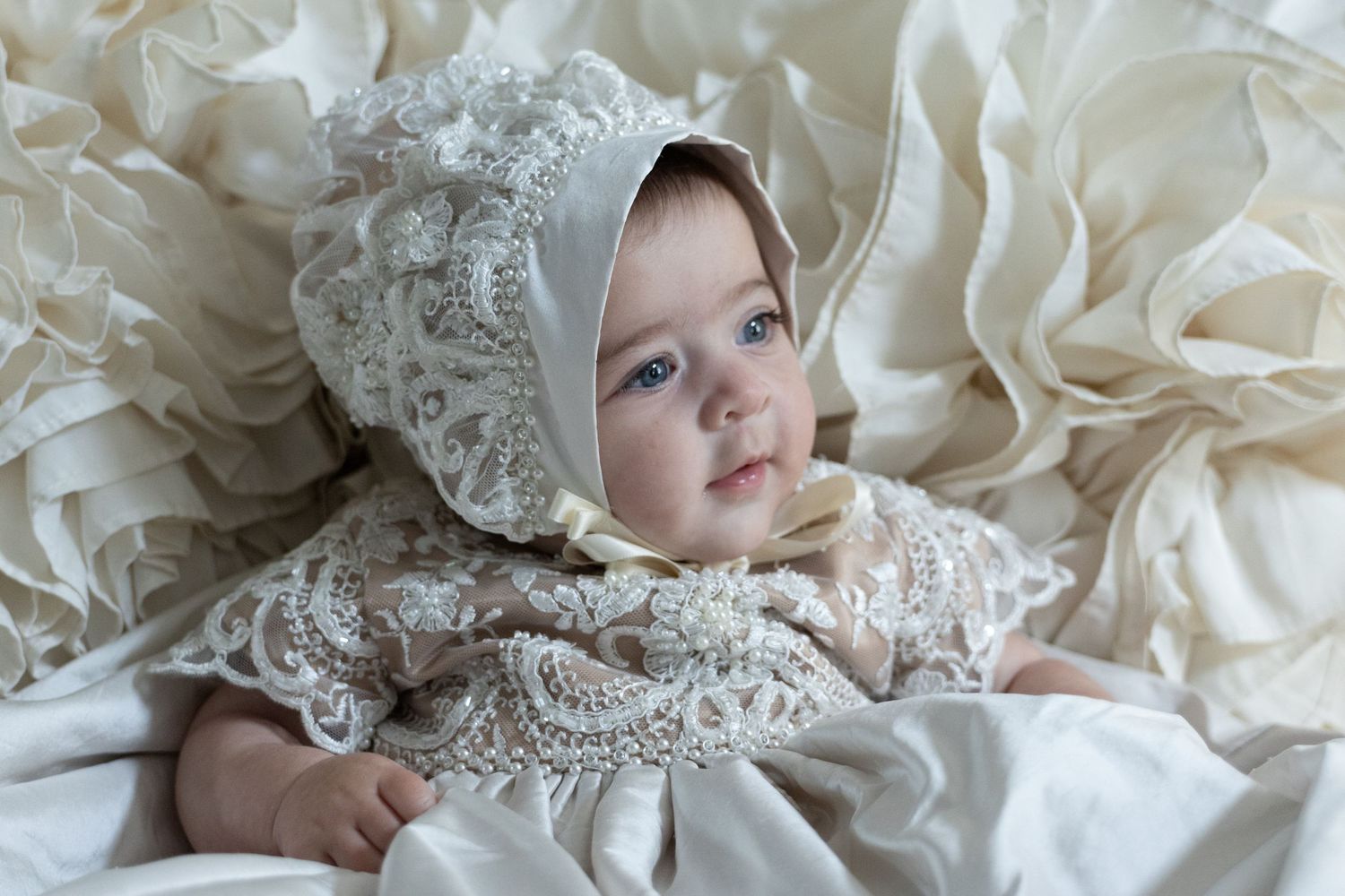 Adorable baby wearing ornate lace christening bonnet and gown lying on ruffled white fabric.