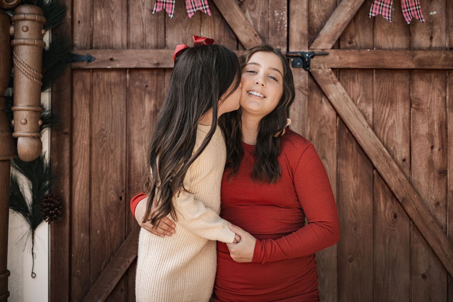 A young girl in a cream dress gives her mother a kiss on the cheek near a rustic wooden barn door.