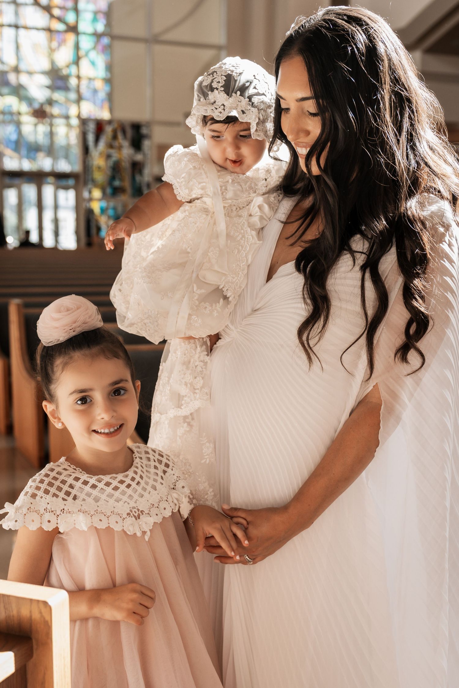 A mother in white dress holds her baby in a lace baptism gown beside her young daughter in a church.