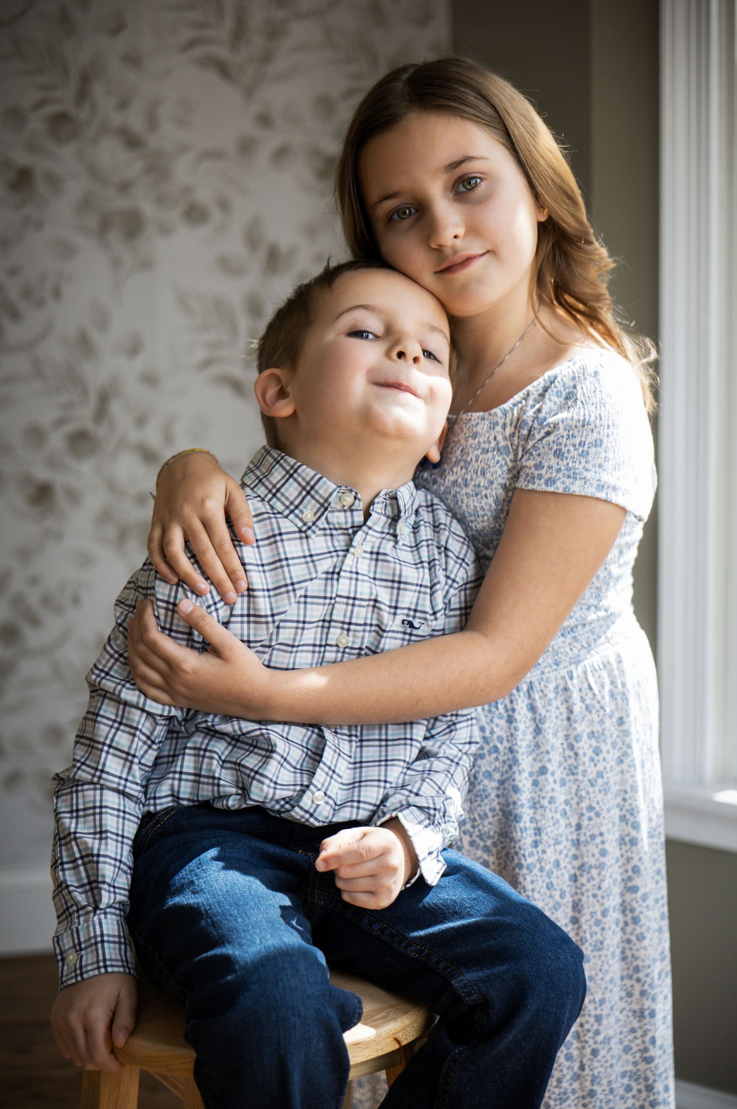 Young girl tenderly hugging her little brother while posing for a portrait near a window in New Jersey.