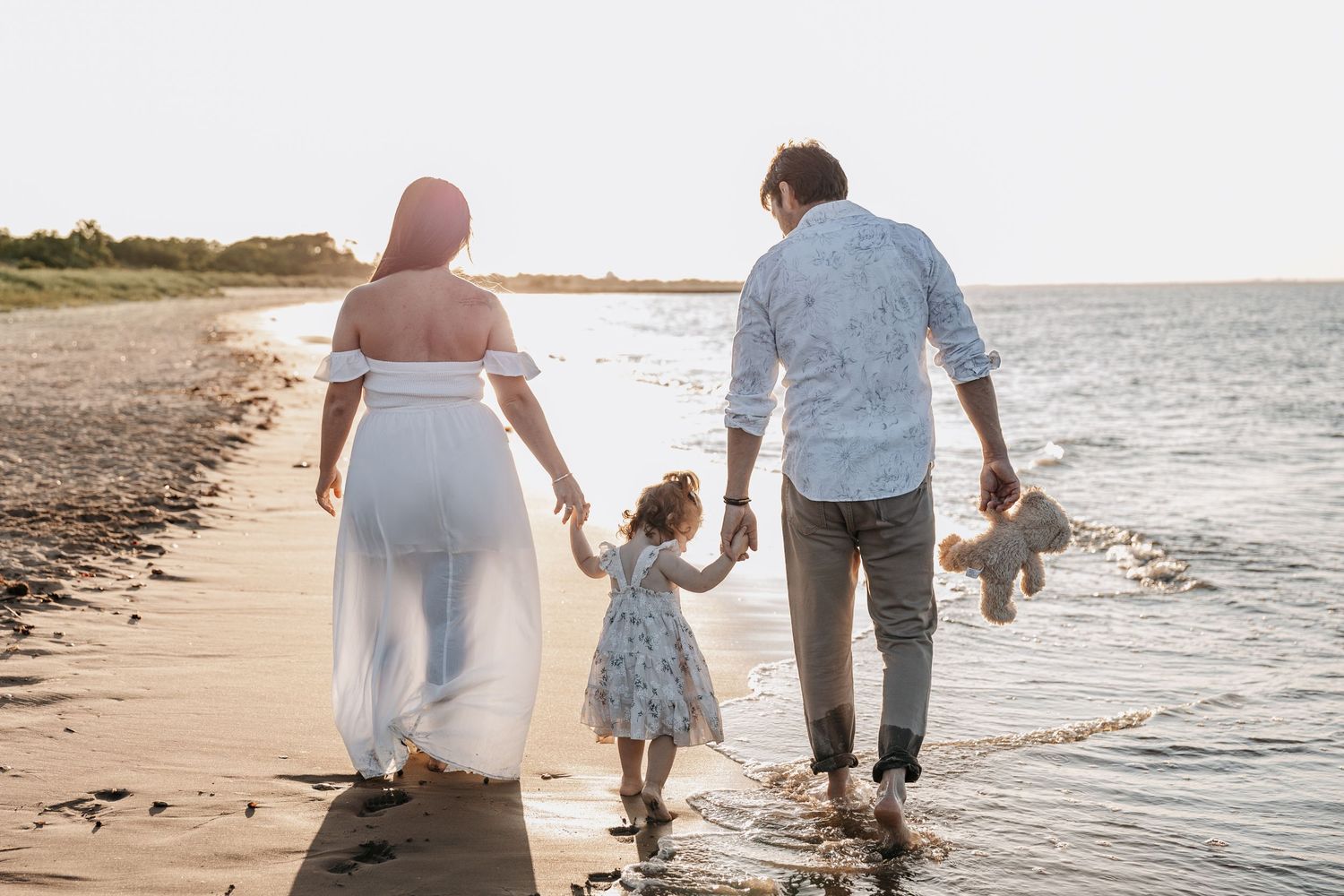 Pregnant woman in white dress walking on beach at sunset with family in New Jersey.