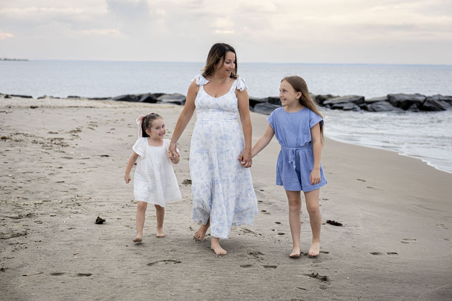 A pregnant mother walks on the beach holding hands with her two young daughters in matching outfits.