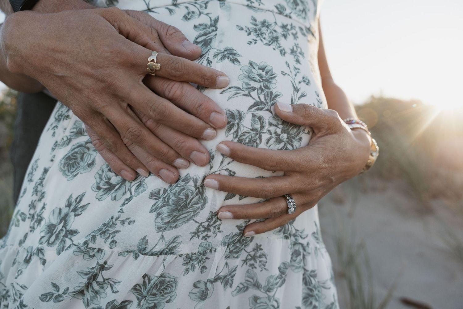 Couple cradling pregnant belly together at sunset, wearing rings in New Jersey.