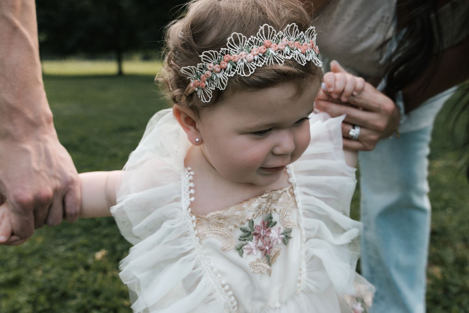 Little girl in white floral dress and tiara headband holds parents hands in park.