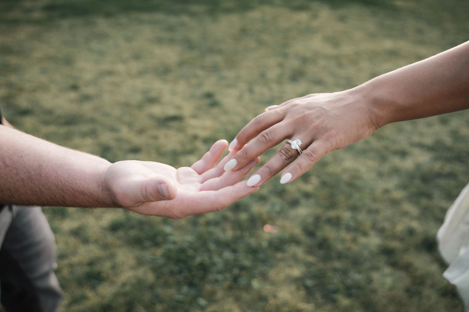 Close-up of bride and groom gently touching hands on grass showcasing diamond engagement ring in New Jersey.