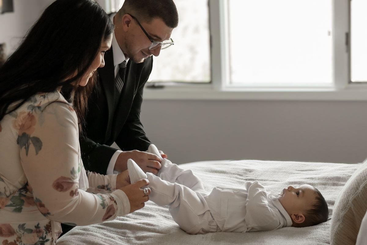 Parents dressing their newborn baby in white christening outfit on a bed.