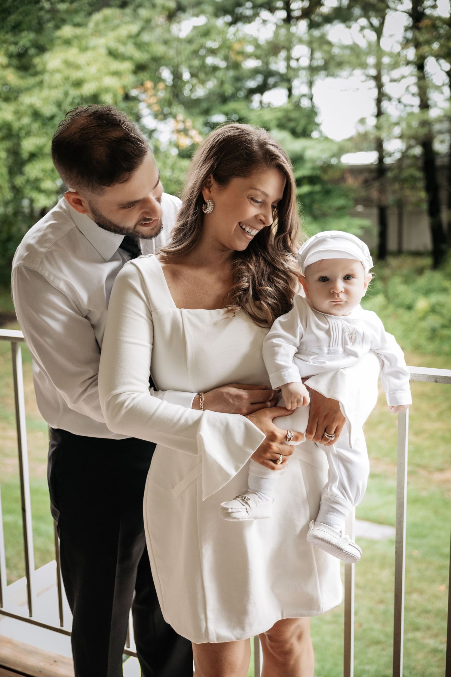 Elegant family portrait with parents in white and black attire holding baby in white christening outfit on balcony.